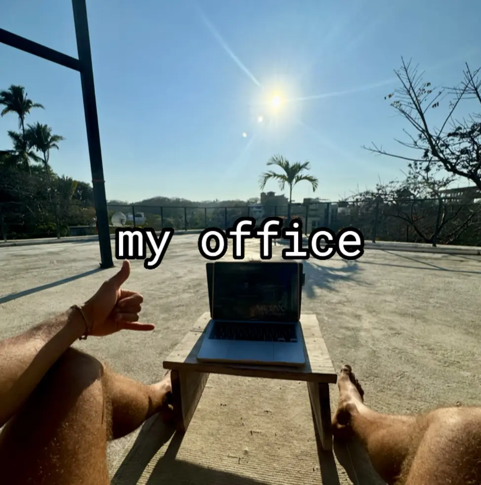 A laptop on a low table beside a pool under palm trees and a bright sun, captioned “my office”